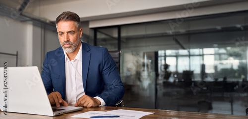 Mature Indian or Latin business man ceo trader using computer, typing, working in modern office doing online data market analysis, thinking planning tech strategy looking at laptop. Banner, copy space