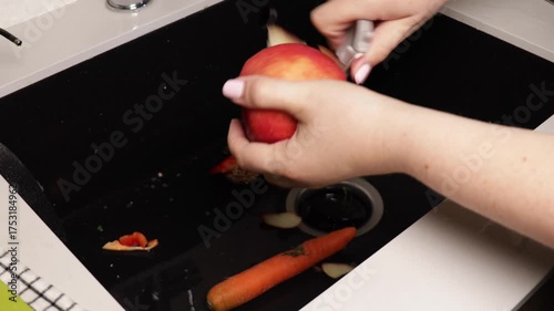peeling apple over sink with food waste shredder, close up of hands removing fruit skin under kitchen faucet, concept of home cooking, waste disposal, kitchen hygiene, household appliance industry