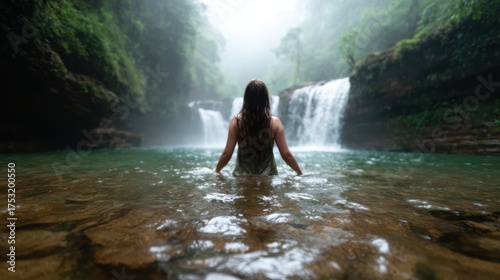 A woman stands in shallow water, enjoying the tranquility of a misty waterfall surrounded by lush greenery, encapsulating the essence of relaxation and nature’s beauty.