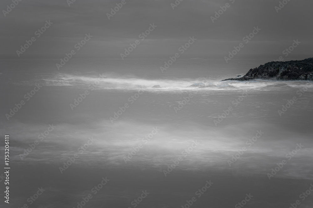 Fototapeta premium Black and white minimalist seascape, smooth water flows past jagged headland with ethereal, misty long-exposure.