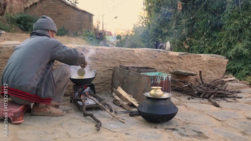 An older Indian man cooks on a small outdoor stove beside a crackling fire in winter. Logs, kettles, and smoke create a rustic village atmosphere, reflecting simplicity, warmth, and daily rural life.