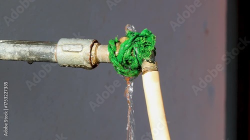 A close-up view of a leaking metal pipe joint wrapped with a bright green rope, dripping water that reflects an improvised plumbing repair and the issue of water scarcity in Indian villages.