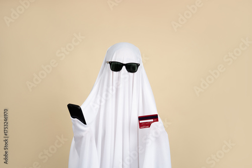 Halloween concept: White ghost wearing black glasses holding a credit card and smartphone on a beige background.