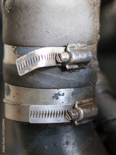 Fotografie Close-up of two metallic hose clamps securing a black rubber or reinforced textile hose, showing wear and tear on the industrial connection