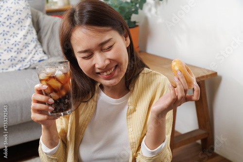 Happy young Asian woman enjoying a sweet glazed donut and iced cola at home, happy smiling in relaxation, showing simple joy and comfort food pleasure in a cozy lifestyle setting
