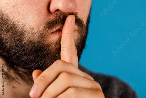 Obraz na plátně Close-up of a bearded man indicating silence by placing his finger on his lips against a blue background, shushing others