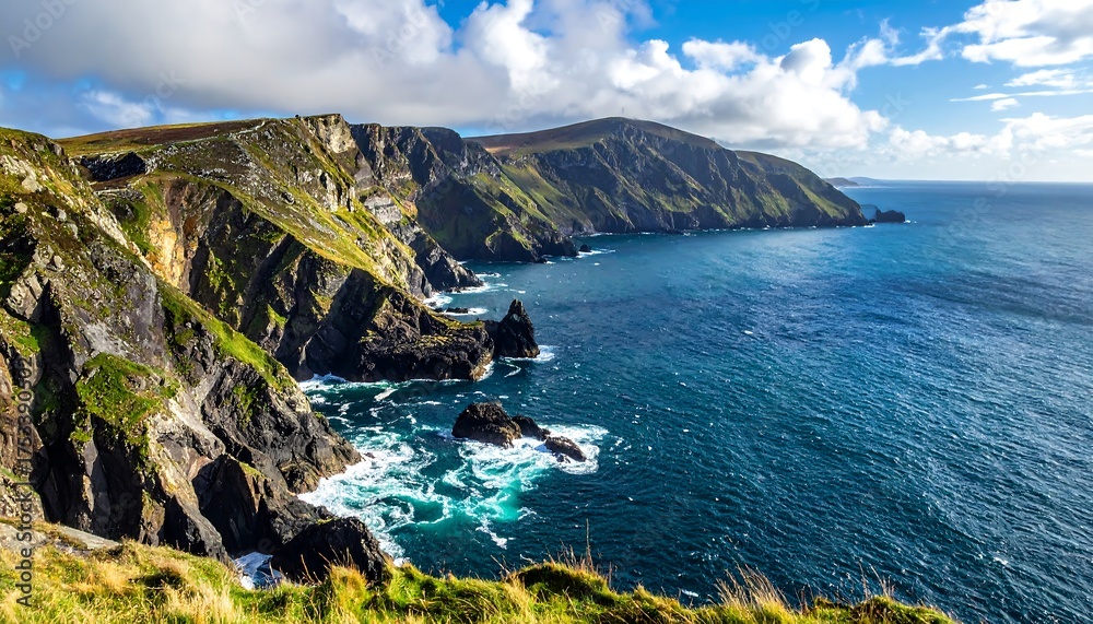 Fototapeta premium A scenic coastal view of a rugged, green cliffside meeting the vibrant blue ocean under a partly cloudy sky. Waves crash against the rocky shore