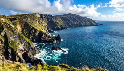 A scenic coastal view of a rugged, green cliffside meeting the vibrant blue ocean under a partly cloudy sky. Waves crash against the rocky shore
