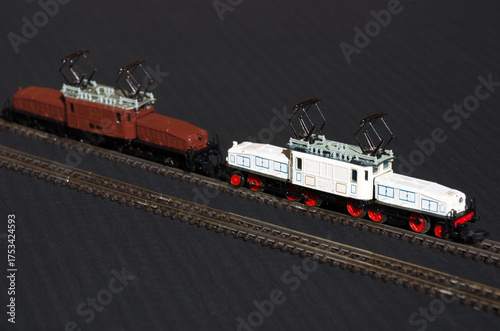 Models of two electric locomotives on miniature tracks against a dark background. The lead train is white with blue accents and red wheels.