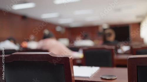 Wooden courtroom chair in focus with blurred people attending a legal trial, symbolizing law and justice.