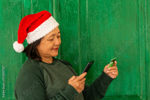 An adult woman, wearing a green sweater and a red Santa Claus hat, is making a transaction using a mobile phone. A green credit card. A green background.