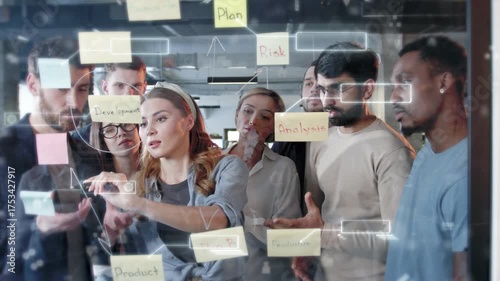 Diverse team of professionals brainstorming around glass board with sticky notes labeled plan, analysis, and product. Holographic diagrams and digital interface overlays showing strategy visualization