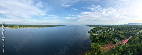 Panoramic aerial view peaceful lakeside village bordered by lush green forests under a clear blue sky, Themes of nature tranquility and rural living.