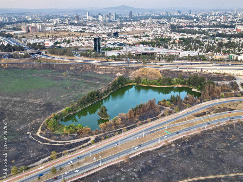 Fototapeta premium Drone photo showing a small lake surrounded by trees and burnt grass near the road to Akron Stadium in Zapopan. The image captures the natural element amidst a dry urban landscape with distant views o
