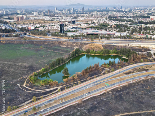 Drone photo showing a small lake surrounded by trees and burnt grass near the road to Akron Stadium in Zapopan. The image captures the natural element amidst a dry urban landscape with distant views o