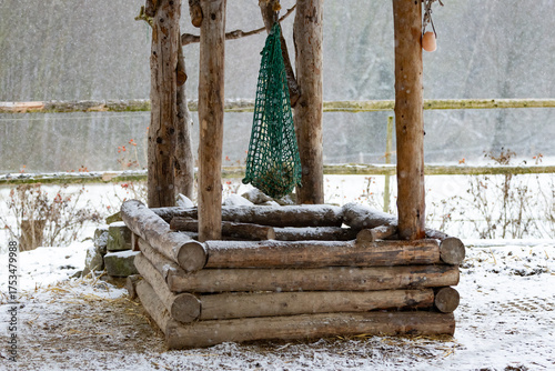 ein grünes fast leeres Heunetz hängt über einer Holz Raufe aus Lärchenholz Baumstämmen an einem Wintertag und es schneit, oben am Balken hängt ein rose Himalaya Salzleckstein
