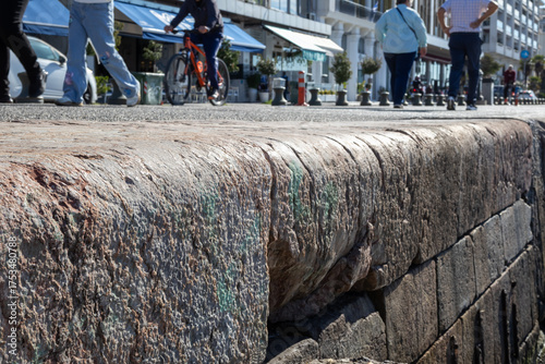 extremely close-up of rough granite seawall or pier edge with blurred pedestrians and cyclist in background on a sunny day.