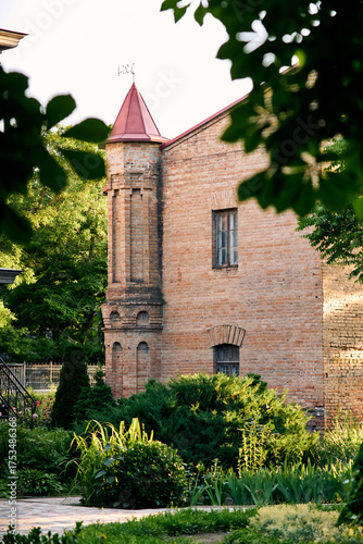 Old brick building with tower and red roof surrounded by green garden plants