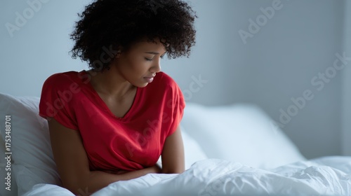 Young African-American woman in bed, feeling unwell and clutching belly due to menstrual pain. Concept of period cramps, abdominal discomfort, and female health