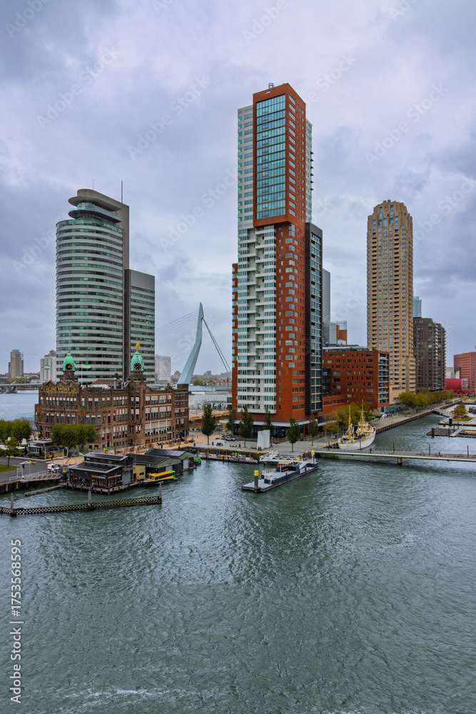 Fototapeta premium The dramatic waterfront skyline of Kop van Zuid in Rotterdam, featuring modern towers, historical buildings, and the Erasmus Bridge across the cloudy harbor water.