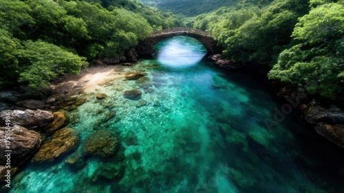 An aerial view of crystal clear water flowing beneath a charming stone bridge, nestled in lush greenery, evoking feelings of tranquility and natural beauty.