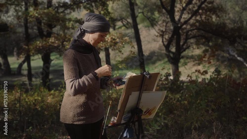 Artist in headscarf painting landscape with watercolor of autumn park on sunny day