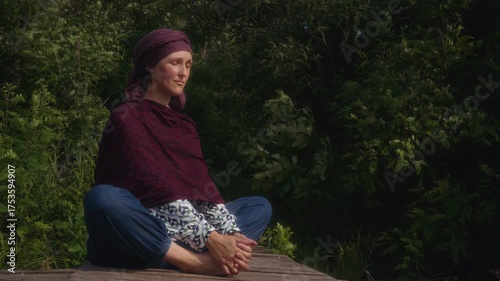 A yogini in a headscarf with her eyes closed is sitting in meditation on a wooden pier by the river
