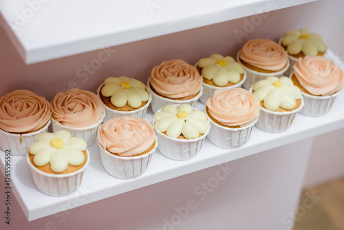 A neat row of custom-made vanilla cupcakes with pale pink rose and creamy yellow floral frosting arranged on a white shelf, perfect for a subtle and elegant celebration or wedding