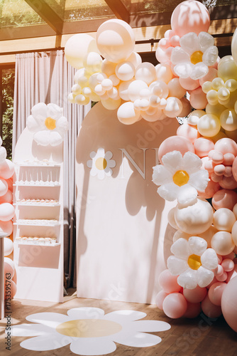 A stylish and sunny first birthday party photo zone with a charming daisy theme, featuring a pastel pink and yellow balloon arch and a candy bar