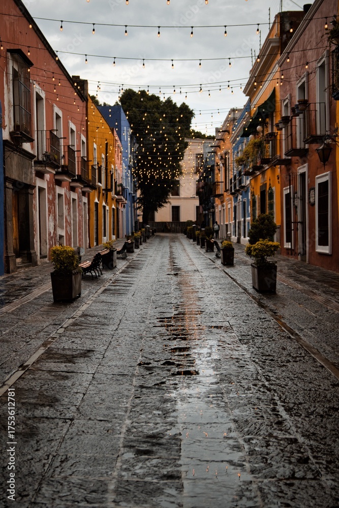 Fototapeta premium Vibrant nighttime scene of a picturesque, rain-soaked alleyway in a historic European town, illuminated by twinkling string lights and warm building colors.