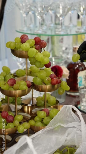 A decorator in black gloves places green and red grapes in a composition in a golden bowl for a wedding table.