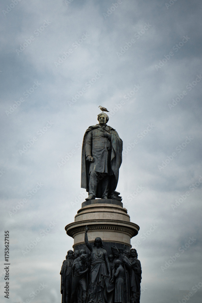 Fototapeta premium Ominous statue of a historical figure stands tall against a moody, cloudy sky, with a lone bird perched atop.