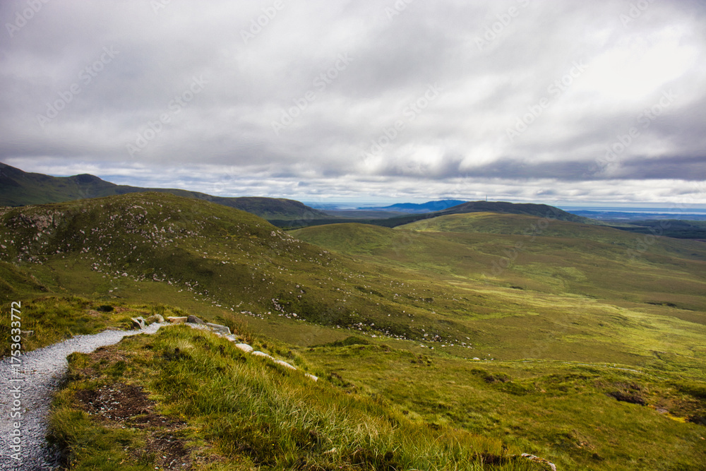 Fototapeta premium Scenic mountainous landscape under dramatic cloudy skies, with a grassy path winding through the rolling hills.