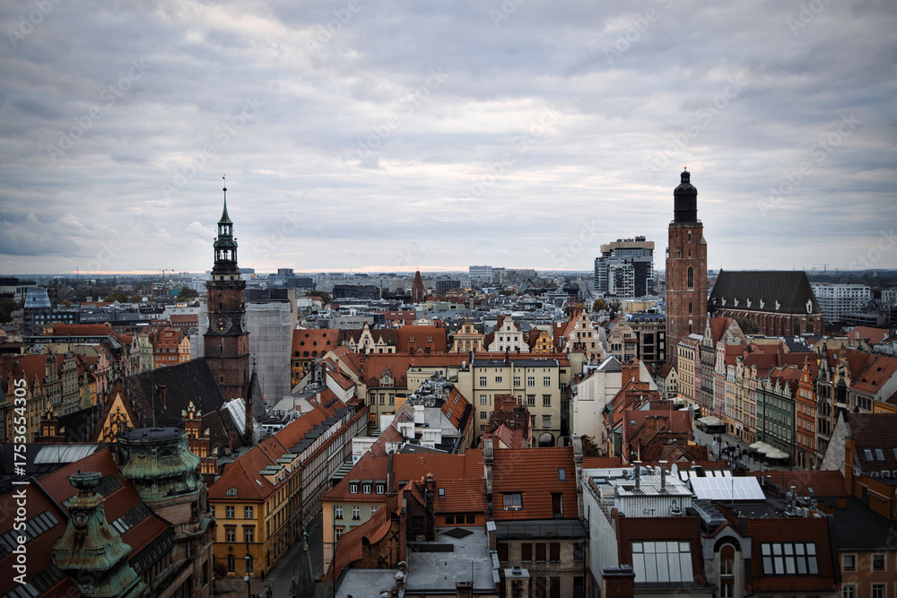 Fototapeta premium Sweeping aerial view of the historic cityscape of Wroclaw, Poland, with its iconic red-roofed buildings, ornate church spires, and modern high-rises on a cloudy day.