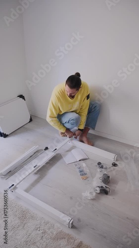 A man in a lemon-colored sweatshirt and jeans sits on the floor, carefully reviewing the instruction manual while sorting metal and wooden parts for an adjustable home office desk. Tools and bolts are