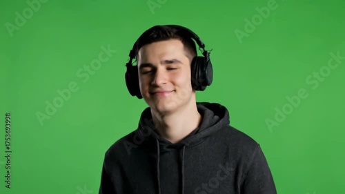 Content Young Man in Hoodie Enjoying Music on Headphones Against Green Screen.
