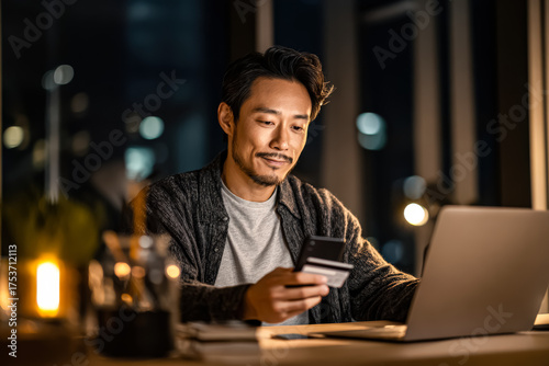 Male individual is seated at a desk, using smartphone and laptop, surrounded by warm lighting, reflecting a contemporary lifestyle and digital engagement