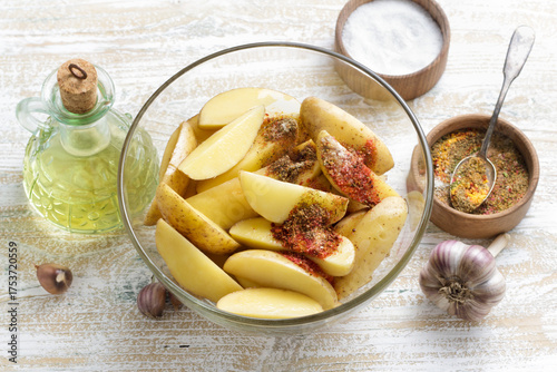Fresh raw potato wedges in glass bowl with garlic, vegetable oil and colorful spices on rustic wooden table, cooking ingredients