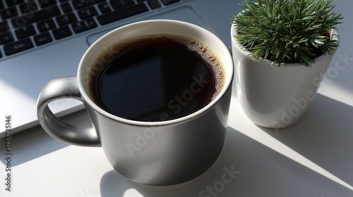 Black coffee in gray mug next to green plant and open laptop on sunny desk