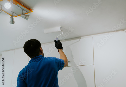 A young oriental man paints the ceiling near the window with a roller.