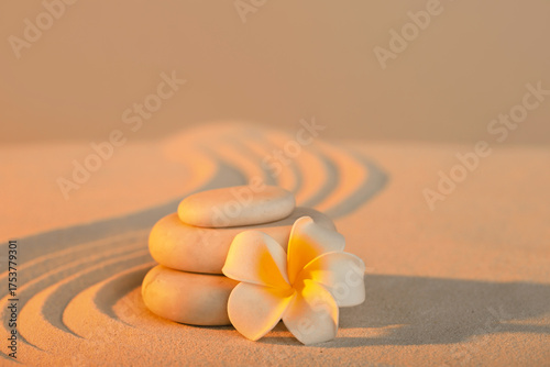 Canvas Print Stack of spa stones with plumeria flower on light sand at sunset