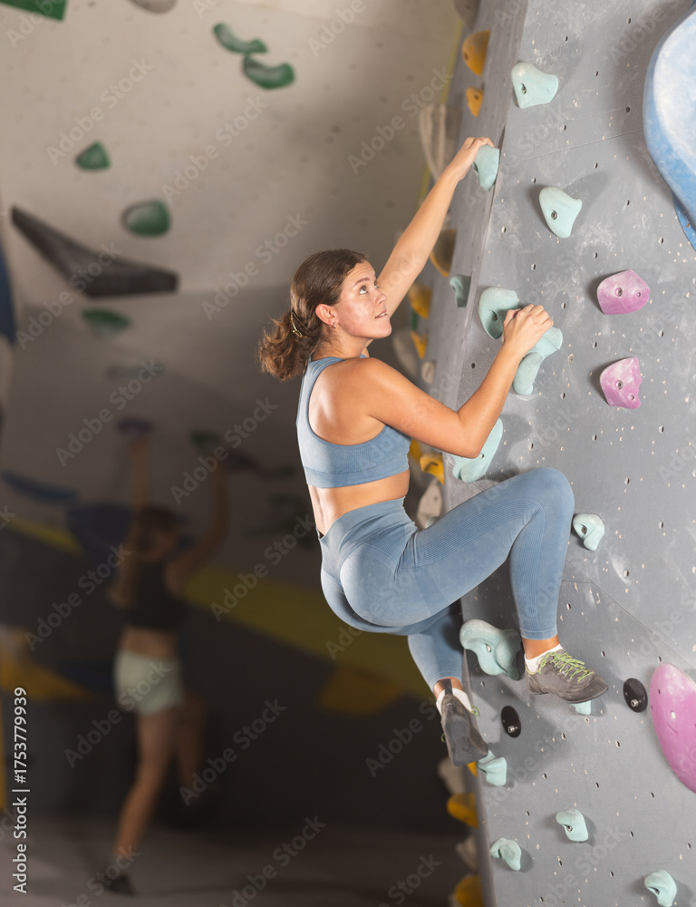 Fototapeta premium Young female athlete in tights, climbs steep cliff artificial wall in gym, physical exertion, struggle with physical inactivity. Universal sports, individual training, curation.
