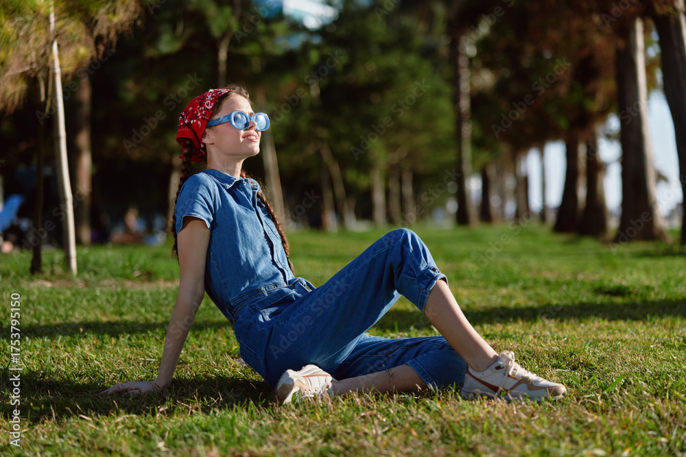 Fototapeta premium lifestyle woman in modern boho-western denim street style relaxes outdoors on grass with film color effect, wearing sunglasses and red headscarf on sunny day