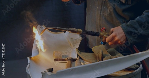 Shaping metal panel metalworker wearing jacket using blowtorch on drum in shop, with hammers mallet