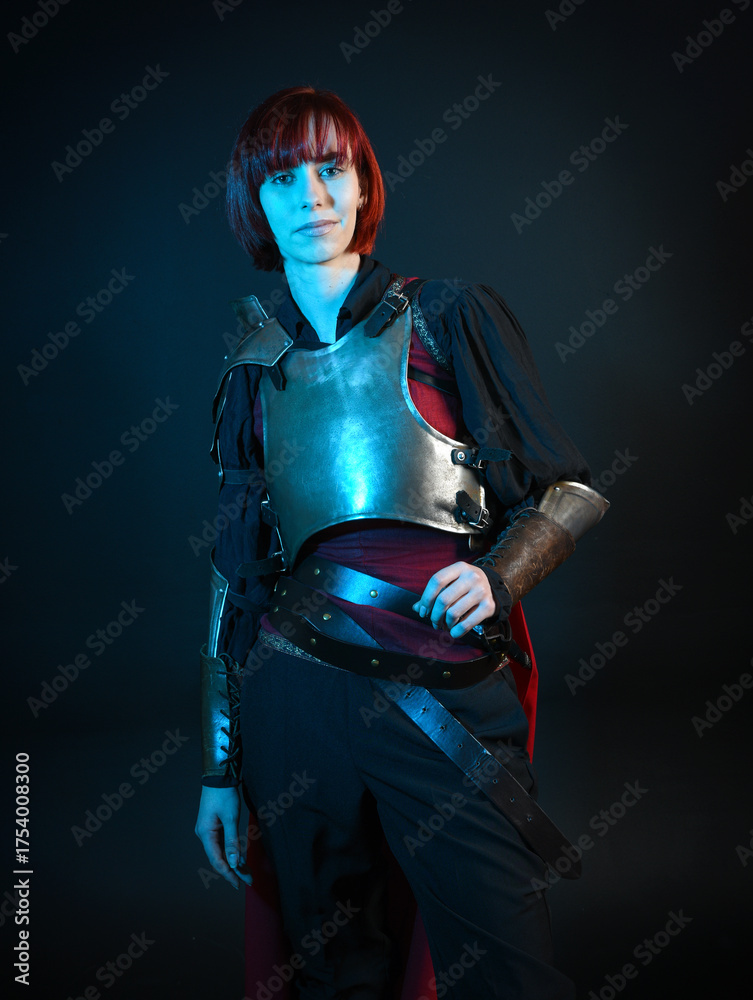 Naklejka premium Fantasy warrior woman model in historical medieval armour costume, red fairytale cloak, cinematic portrait of strong fantasy knight holding sword. Epic pose, silhouette dark studio background