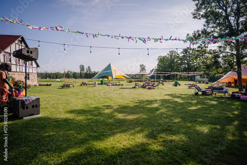 Fotografie Colorful outdoor hippie-style festival setup with picnic benches, decorated tents, string lights, and festive ribbons on a sunny day in a grassy field