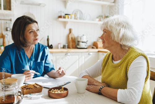 Elderly woman with grey hair answering questions of female social worker sitting at kitchen table drinking tea as first acquaintance before providing caregiver support program for aged people