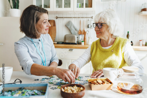 Senior lady in glasses solving puzzle with assistance of her caregiver sitting at kitchen table, having warm friendly relationships during her alzheimer rehabilitation therapy at nursing home
