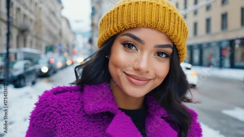 A young woman wearing a bright yellow hat and a purple coat smiles at the camera, surrounded by the city atmosphere.