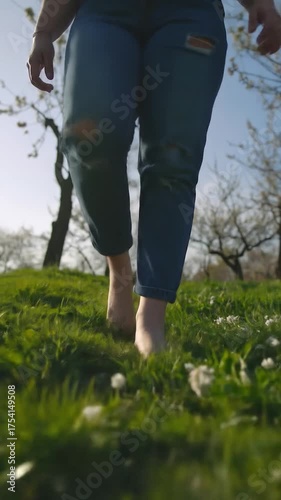 A person in ripped jeans walks barefoot through lush green grass with trees in the background, enjoying nature